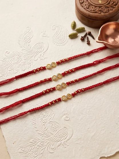 Four red braided Rakhi strings with gold-toned decorative beads and small metallic accents, arranged diagonally on a textured off-white paper with embossed peacock motifs. Decorative props include a copper diya, cardamom pods, cloves, and a carved wooden box in the upper right.