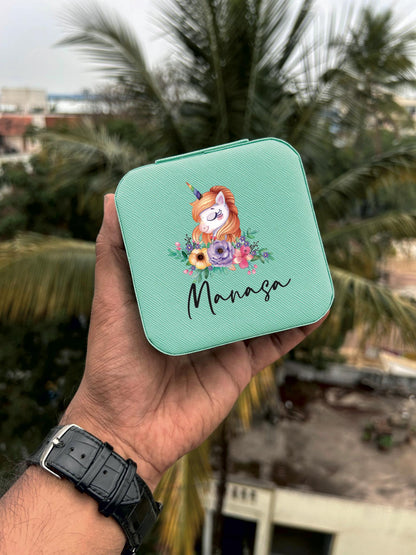 A personalized mini jewelry box held in a hand, with a green background and a custom name 'Manasa' on the top. The box features a cartoonish animal illustration.