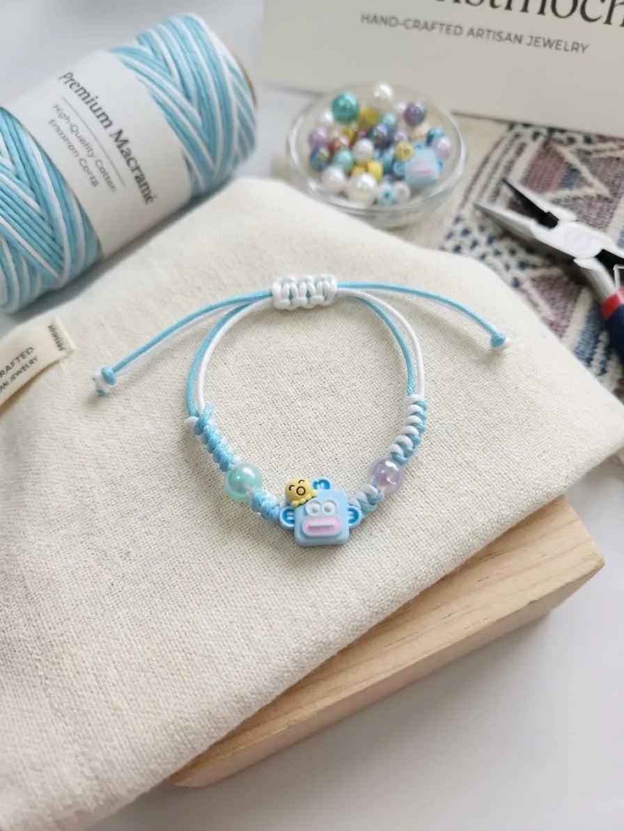 A handmade beaded bracelet with light blue cord, white beads, pastel beads, and a small cartoon character charm, displayed on a cream fabric surface with yarn, beads, and pliers in the background.