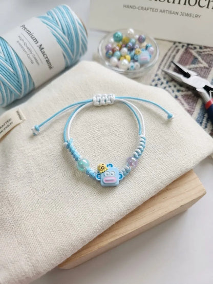 A handmade beaded bracelet with light blue cord, white beads, pastel beads, and a small cartoon character charm, displayed on a cream fabric surface with yarn, beads, and pliers in the background.