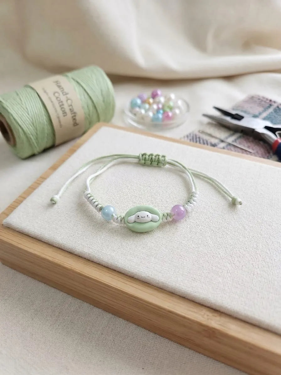A handmade cord bracelet with a mint green central bead featuring a cute face, flanked by pastel blue and lavender beads, displayed on a wooden block with crafting supplies in the background.