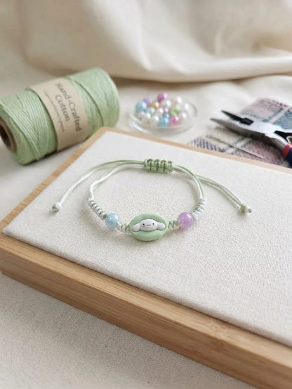 A handmade cord bracelet with a mint green central bead featuring a cute face, flanked by pastel blue and lavender beads, displayed on a wooden block with crafting supplies in the background.