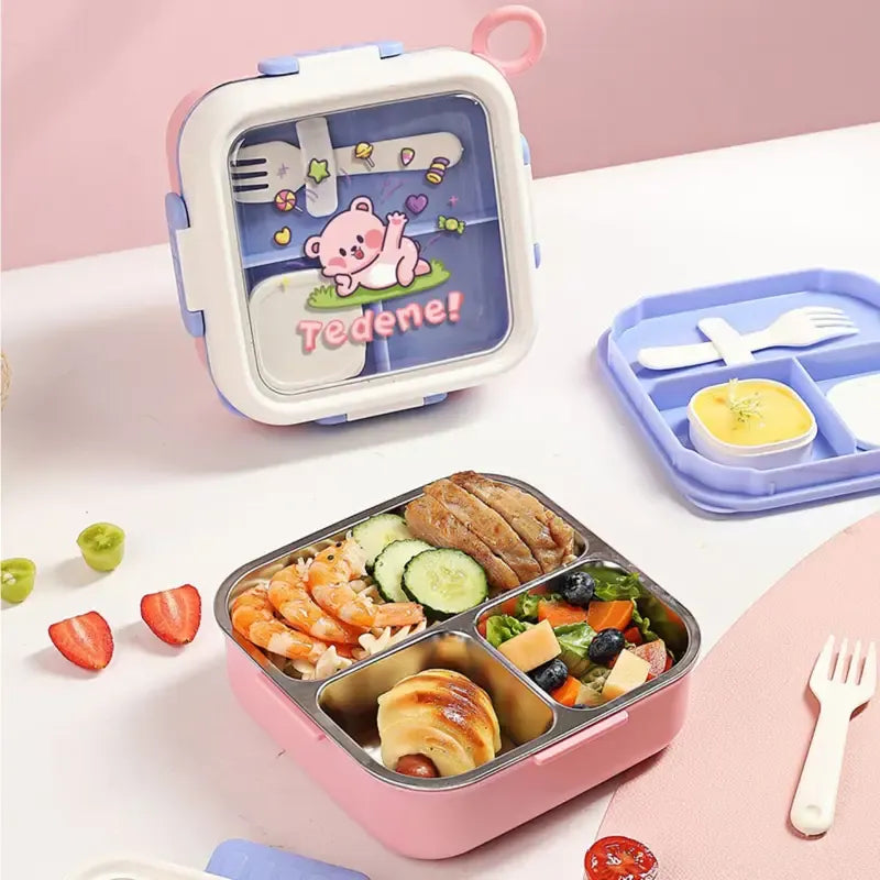 A pink compartment lunch box with a decorative lid featuring a cartoon bear and the text 'Tedene!' is shown open with food inside, alongside a blue tray and utensils on a light pink tabletop.