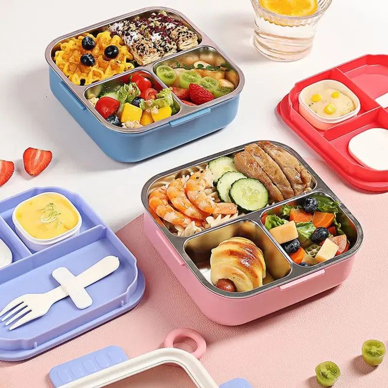 Three colorful compartment lunch boxes in blue, pink, and red are arranged on a pink surface, filled with salads, shrimp, pasta, fruit, and small side dishes. A white plastic fork rests in the blue box, and a glass of water and sliced strawberries are nearby.