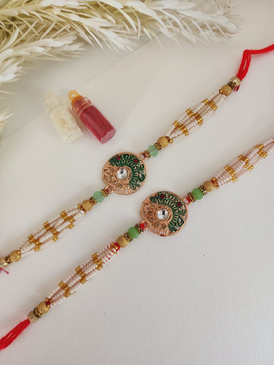 A Hindu woman's rakhi with intricate gold, green, and red beadwork, and a floral design, displayed alongside a small red bottle and a card.