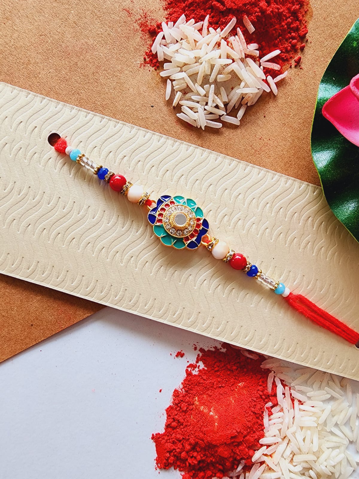 A multicolor traditional Indian Rakhi with intricate design and decorative elements, displayed on a card with scattered rice grains and red powder.
