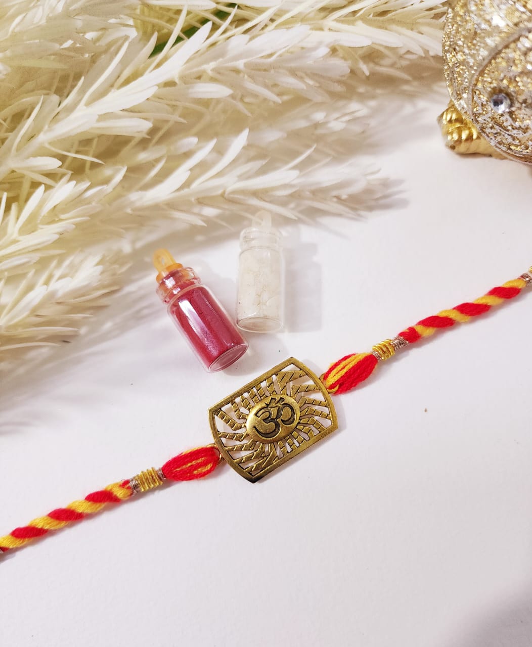 A Hindu 'Rakhi' or bracelet with an om symbol in gold color on a red and yellow thread, displayed alongside small containers of turmeric powder and a perfume bottle.