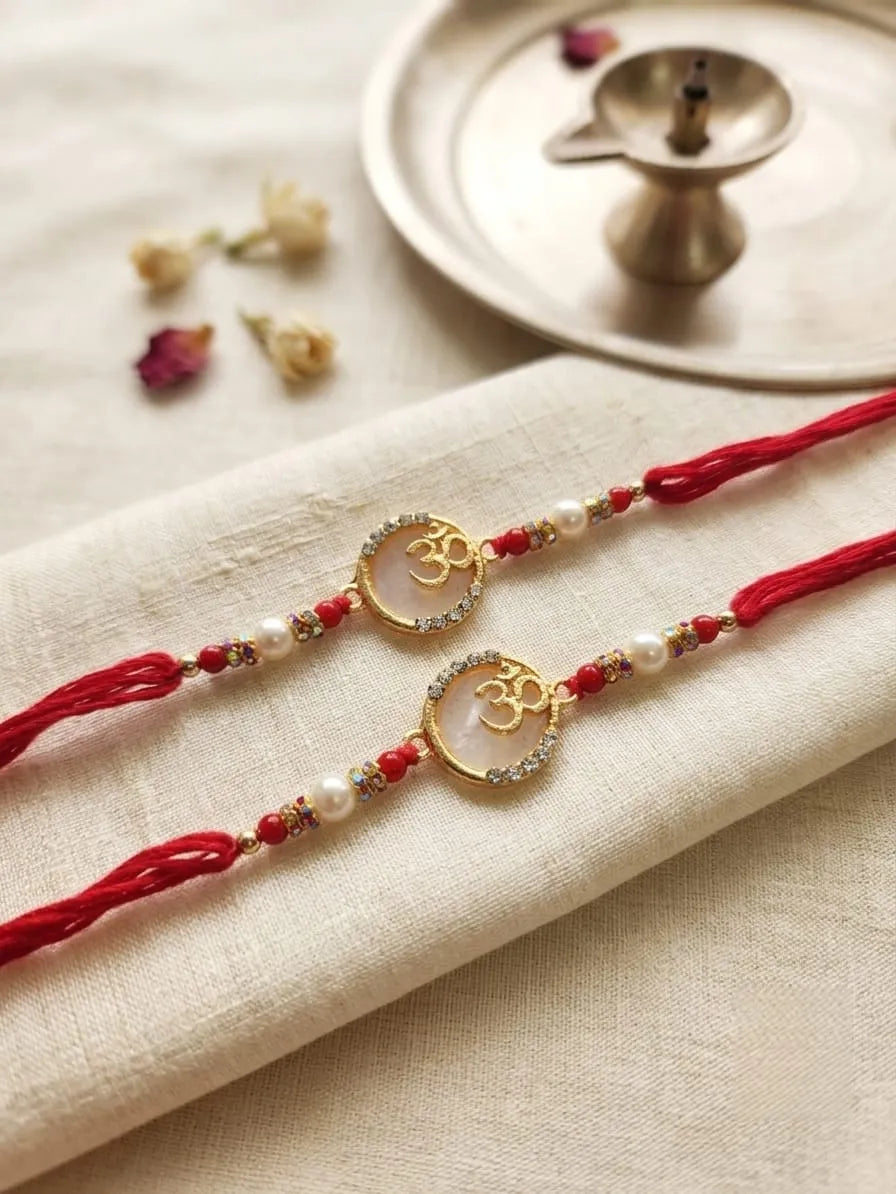 Two red Rakhi strings with gold Om pendants, white pearls, and small red beads, laid on a beige fabric napkin with dried flowers and a brass lamp in the background.
