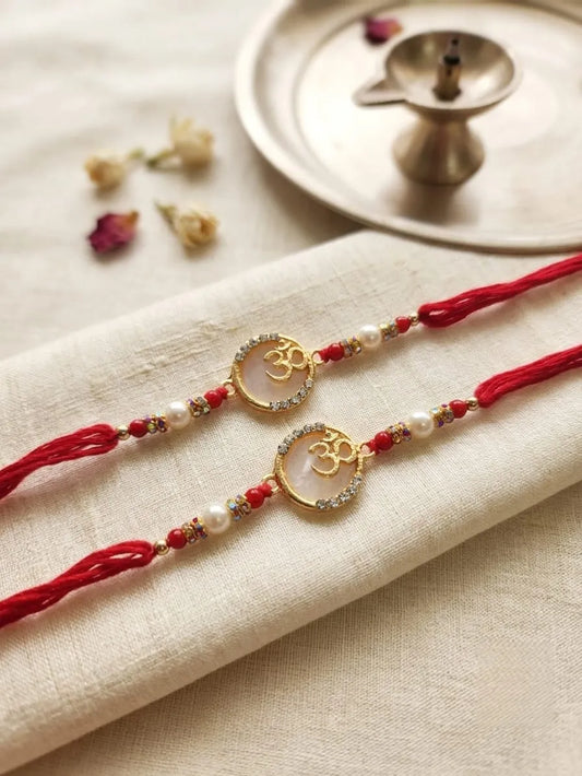 Two red Rakhi strings with gold Om pendants, white pearls, and small red beads, laid on a beige fabric napkin with dried flowers and a brass lamp in the background.