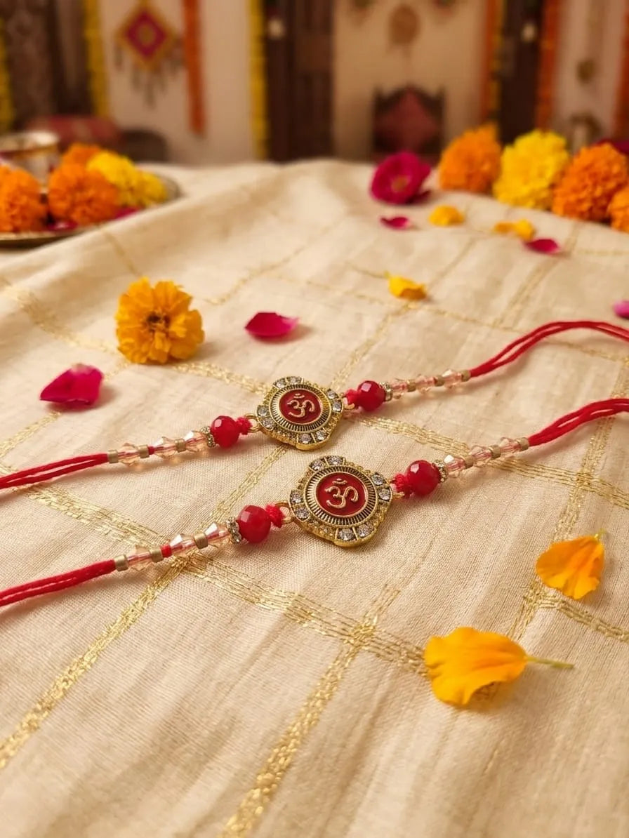 Two traditional Indian Rakhi bracelets with red cords, red beads, and gold-toned Om pendants, displayed on a beige embroidered fabric with marigold flowers scattered around.