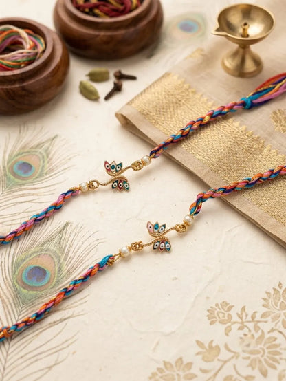 Two colorful braided Rakhi strands with gold-tone owl charms and small pearl beads, arranged on a decorative cream surface with peacock feather, gold fabric, wooden bowls, and a brass diya.