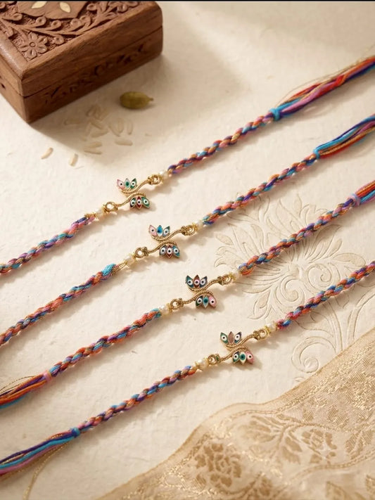 Four colorful braided Rakhi strands with small owl charms, arranged diagonally on a cream-colored textured surface with a carved wooden box in the background.