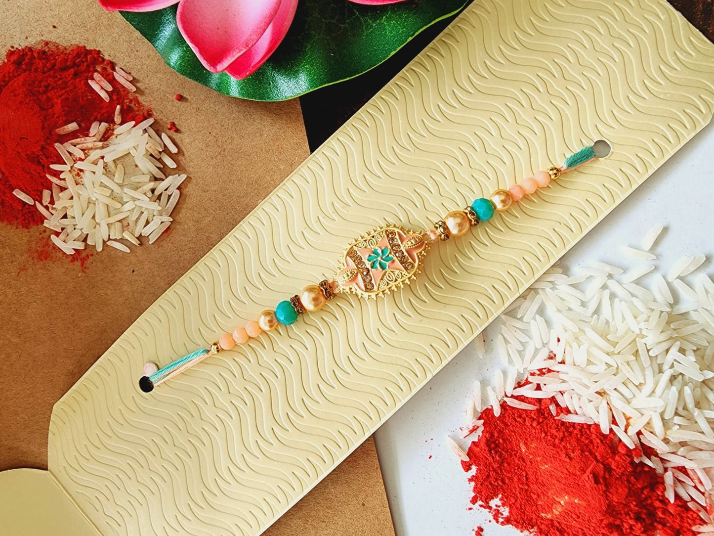 A peach-colored rakhi bracelet displayed on a beige surface with a floral pattern, alongside a small pile of white rice and a red flower for ceremonial purposes.