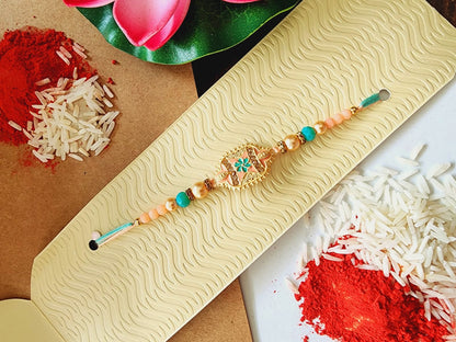 A peach-colored rakhi bracelet displayed on a beige surface with a floral pattern, alongside a small pile of white rice and a red flower for ceremonial purposes.