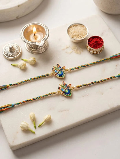 Two braided Rakhi strings with colorful beads and gold-tone bee motifs, arranged on a white marble slab with a lit diya, rice, red powder, and small flower buds.