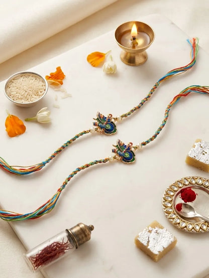 Two traditional Indian braided silk Rakhi bracelets with peacock and floral charms, styled on a white marble surface with festive props including a brass diya, rice, sweets, and flowers.