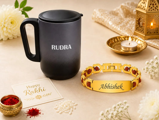 A black insulated mug with the name 'RUDRA' printed on it, displayed alongside a gold-toned bracelet engraved with 'Abhishek' and accented with red stones. The scene is decorated for Rakhi with a 'Happy Rakhi' card, red powder, white flowers, a lit candle, and a brass lantern on a beige surface.