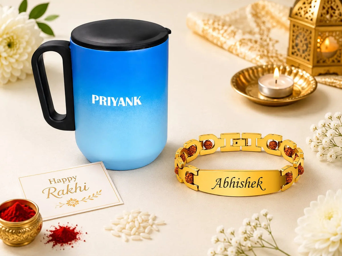 A blue-to-light-blue gradient insulated mug with a black lid and handle, printed with the name 'PRIYANK', displayed alongside a gold-tone engraved bracelet with red stones and the name 'Abhishek'. The scene is styled for Rakhi with white flowers, red powder, a lit diya, and a 'Happy Rakhi' card on a cream background.
