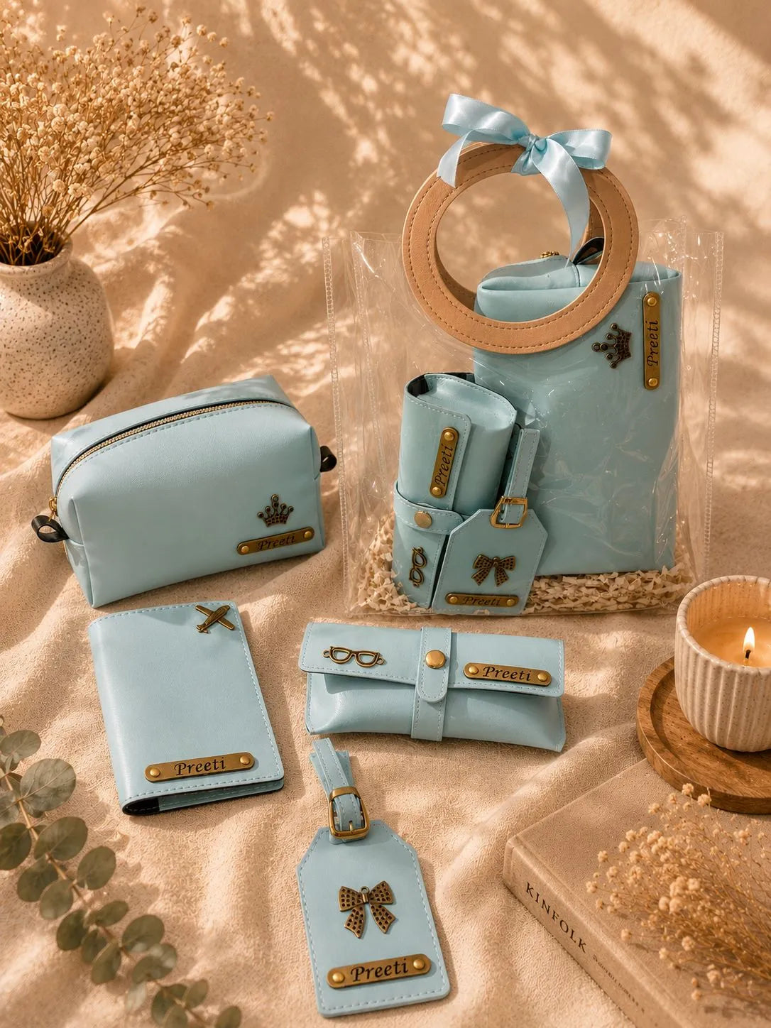 A coordinated set of light blue faux-leather accessories arranged on beige fabric with dried flowers, a candle, and a book. The set includes a small zip pouch, a wallet, a ring holder with a circular handle, a tote bag, a small pouch, a luggage tag, and a card holder, all with gold-tone hardware and bow motifs.