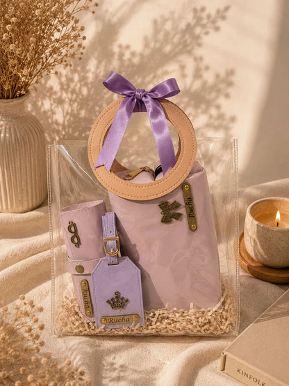 A clear plastic gift bag containing a lavender quilted handbag with a circular tan handle, a matching lavender pouch, and a lavender luggage tag, all tied with a purple satin bow. The set is styled on a beige fabric surface with dried flowers, a candle, and a box in a warm, sunlit setting.