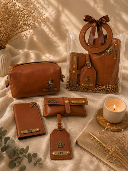 A coordinated set of brown leather accessories arranged on a cream fabric surface with dried flowers, a candle, and a book. The set includes a zip pouch, a small card holder, a wristlet or strap bag, a luggage tag, and a clear tote bag with a circular handle and bow.