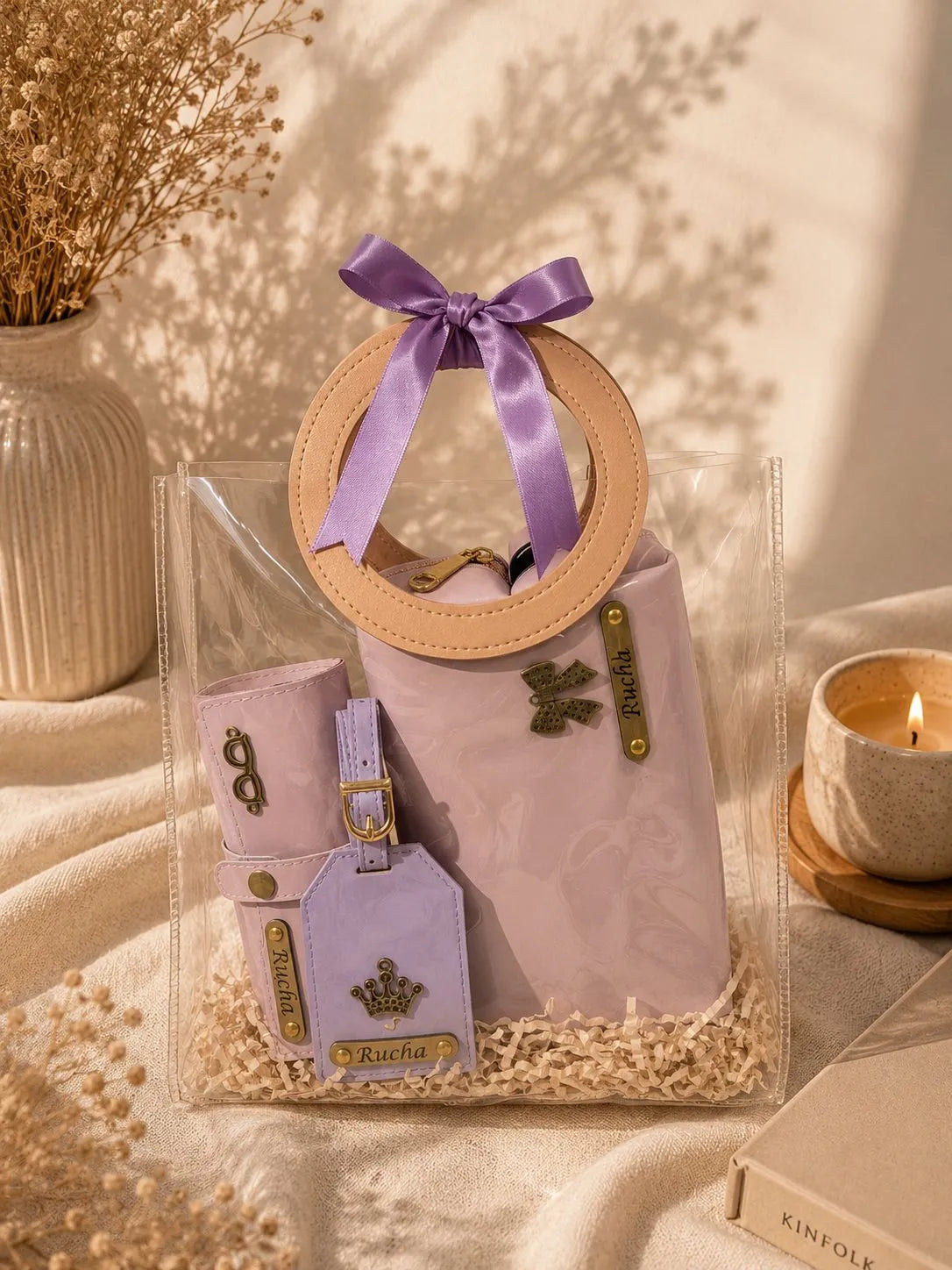 A clear plastic gift bag containing a lavender quilted handbag with a circular tan handle, a matching lavender pouch, and a lavender luggage tag with gold hardware. The bag is tied with a lavender satin ribbon and styled on a beige fabric surface with dried flowers, a candle, and a box in a warm, sunlit setting.