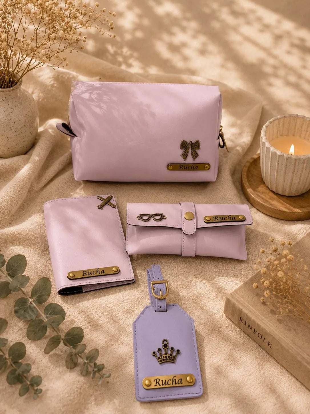 A coordinated set of light pastel pink and lavender leather accessories arranged on beige fabric with dried flowers, a candle, and a book. The set includes a large zip pouch, a slim wallet, a rolled sleeve, a luggage tag, and small hardware accents, all branded Rucha with gold-tone logo plates.