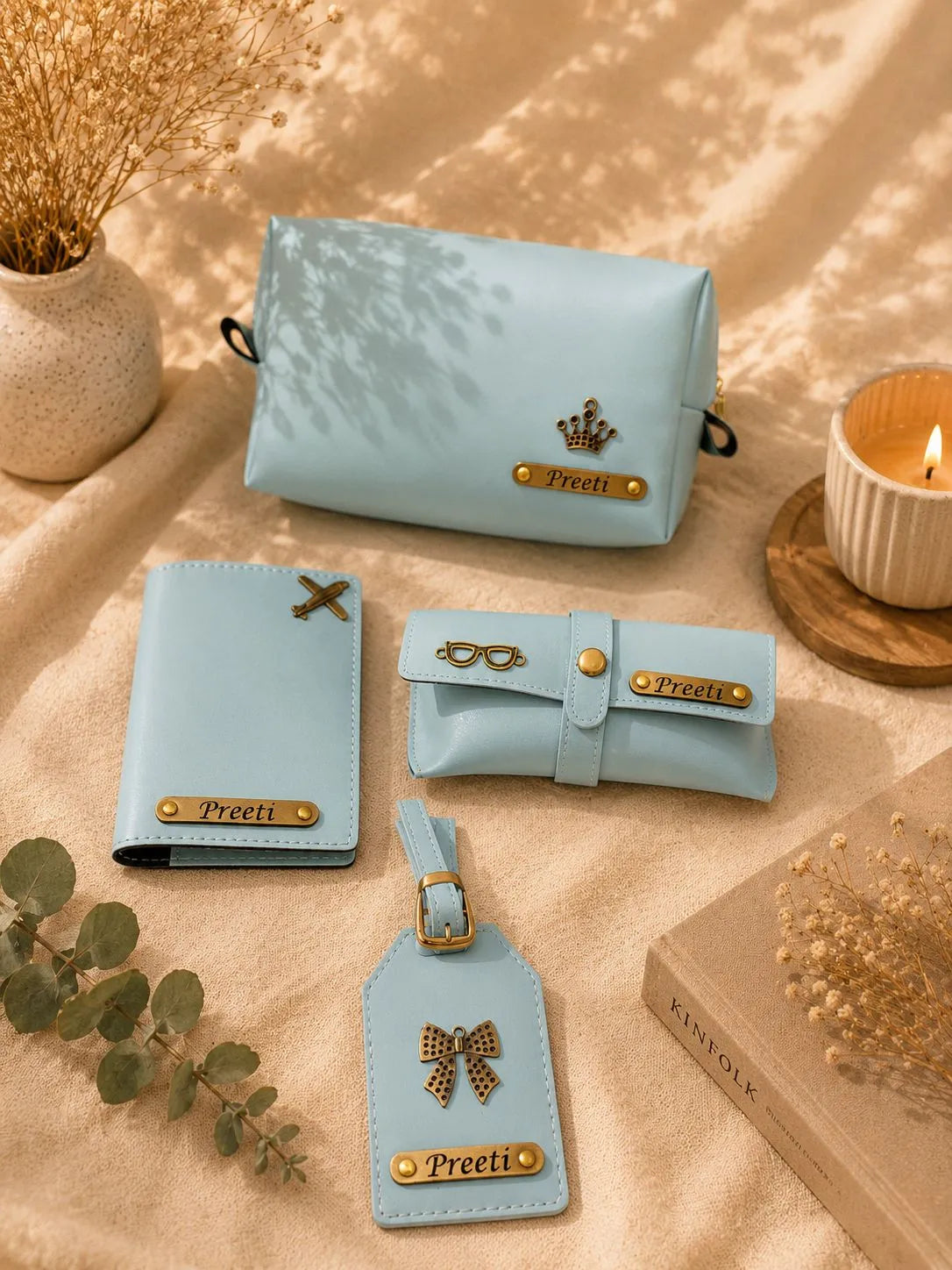 A coordinated set of light blue faux-leather accessories arranged on a beige fabric surface with dried flowers, a candle, and a book. The set includes a zip pouch, a wallet, a rolled sleeve, and a luggage tag, each with gold-tone hardware and a nameplate reading 'Preeti'.