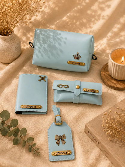 A coordinated set of light blue faux-leather accessories arranged on a beige fabric surface with dried flowers, a candle, and a book. The set includes a zip pouch, a wallet, a rolled sleeve, and a luggage tag, each with gold-tone hardware and a nameplate reading 'Preeti'.