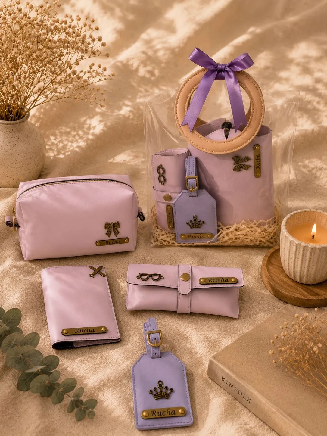 A coordinated pastel pink and lavender luggage set arranged on a beige fabric surface with dried flowers, a candle, and a book. The set includes a tote bag with a wooden ring handle and purple ribbon bow, a zip pouch, a wallet, a small pouch, a luggage tag, and a belt bag, all with gold-tone hardware and decorative bows or crowns.