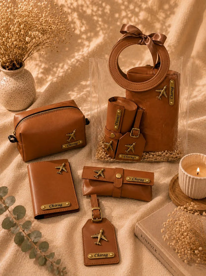 A coordinated set of brown leather travel accessories arranged on a beige fabric backdrop with dried flowers, a candle, and a book. The set includes a small zip pouch, a wallet, a card holder, a luggage tag, a belt, and a tote bag with a circular handle, all featuring small gold-tone airplane motifs and the name 'Chengyu' on the pieces.