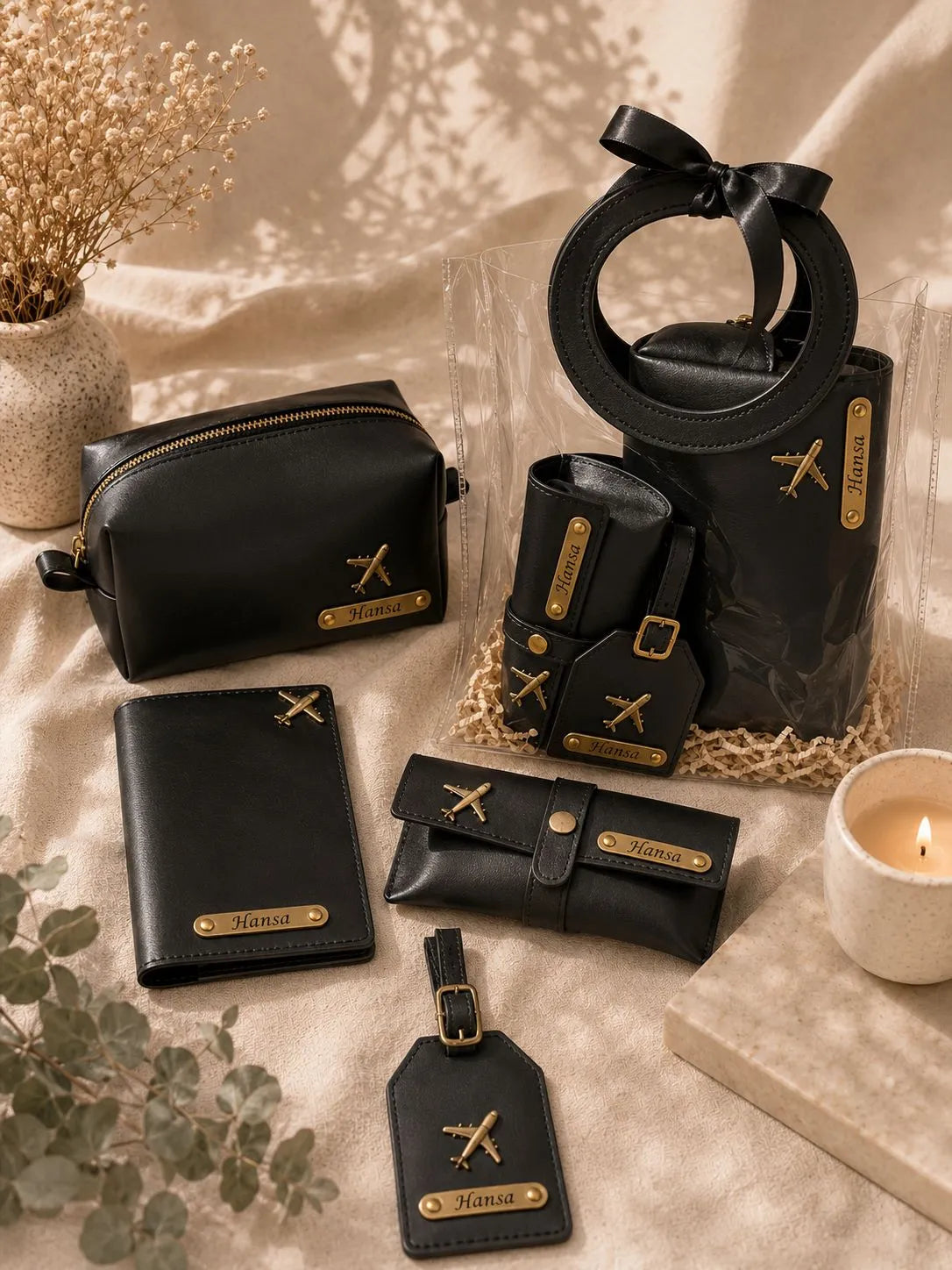 A coordinated set of black leather accessories with gold-tone hardware and logo plates, arranged on beige fabric with dried flowers, a candle, and a clear gift bag. The set includes a large tote bag with a circular handle, a zip pouch, a wallet, a small pouch, a strap, and a luggage tag.