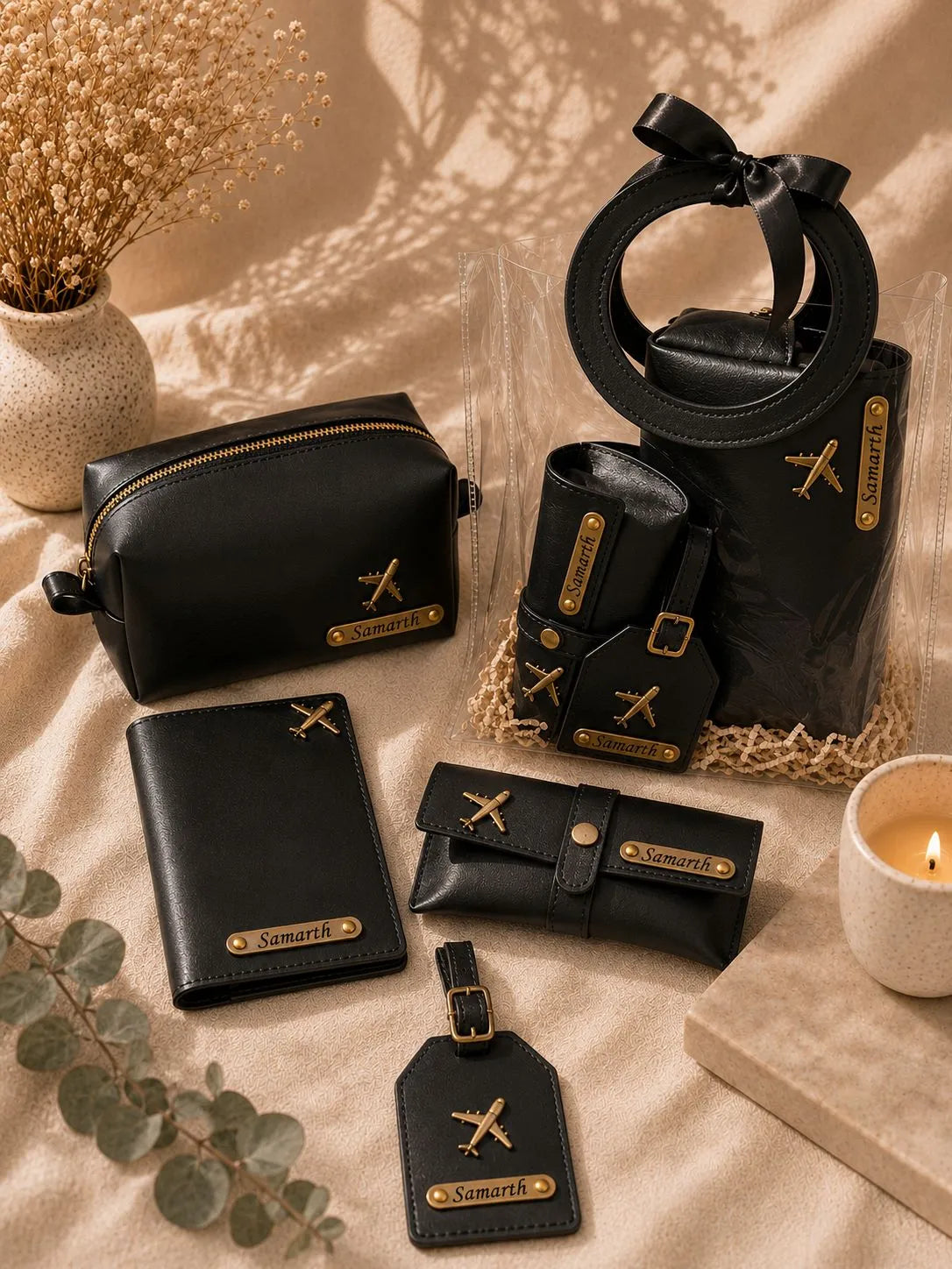A coordinated set of black leather travel accessories with gold-tone hardware and airplane motifs, arranged on a beige fabric surface with dried flowers, a candle, and a small block. The set includes a zip pouch, wallet, card holder, small pouch, tote bag with a circular handle, and luggage tag.
