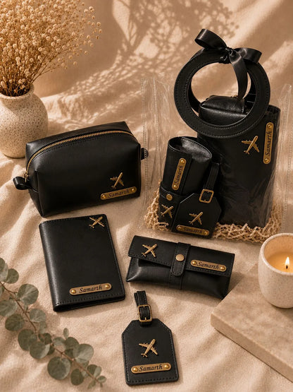A coordinated set of black leather travel accessories with gold-tone hardware and airplane motifs, arranged on a beige fabric surface with dried flowers, a candle, and a small block. The set includes a zip pouch, wallet, card holder, small pouch, tote bag with a circular handle, and luggage tag.
