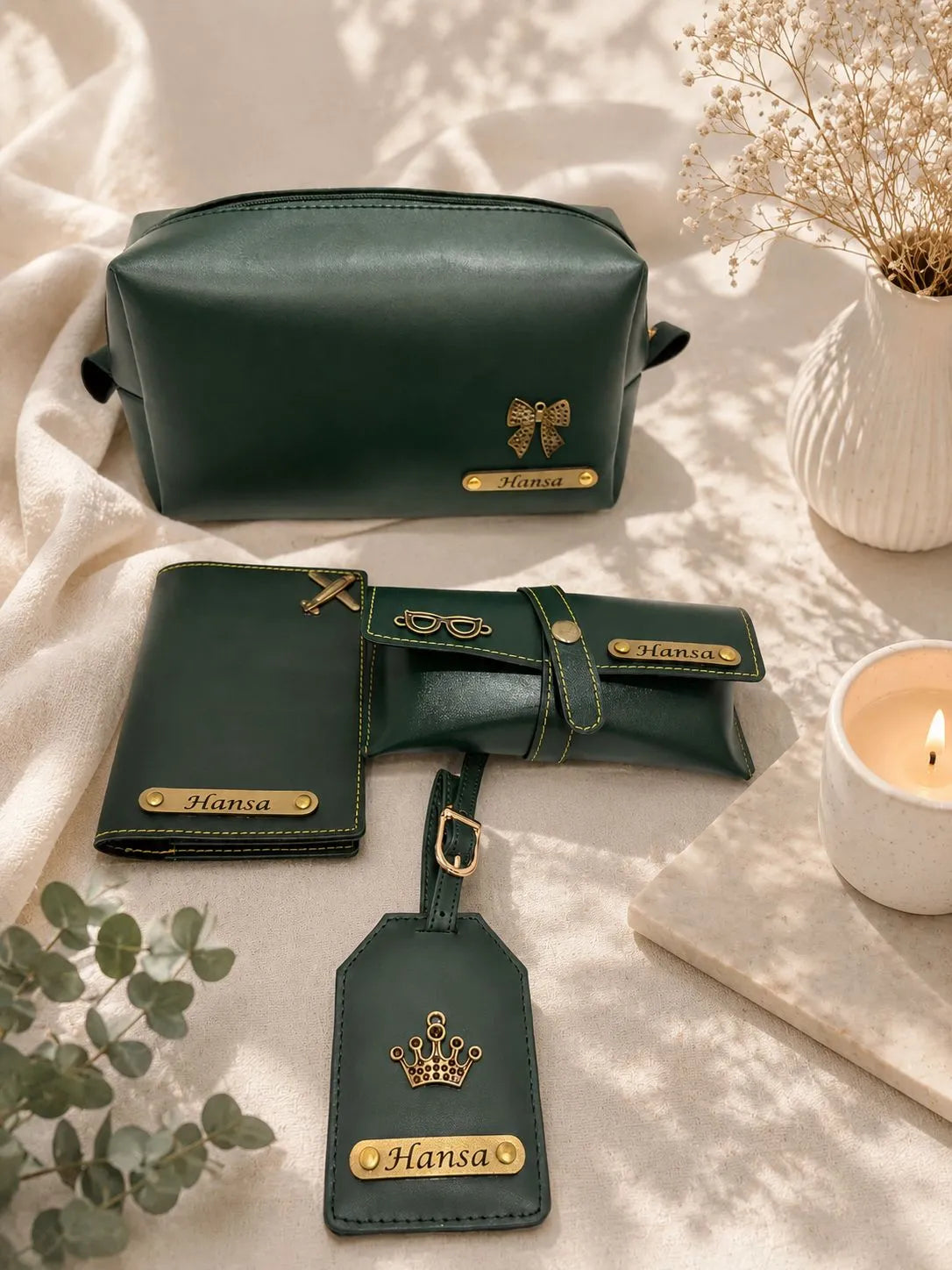 A coordinated dark green leather travel set laid out on a cream fabric surface with soft shadows, including a zip pouch, wallet, passport holder, and luggage tag, each with gold-tone hardware and the name 'Hansa' on a gold plaque.