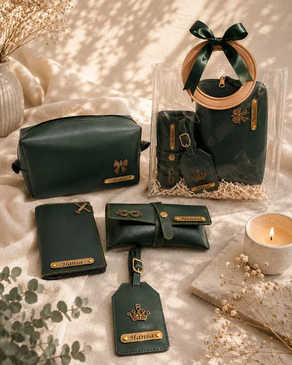 A coordinated set of dark green leather accessories arranged on a beige fabric surface with dried flowers, a candle, and a clear gift bag. The set includes a zip pouch, wallet, card holder, luggage tag, and a small bag with a circular handle, all with gold-tone hardware and a bow on the bag.
