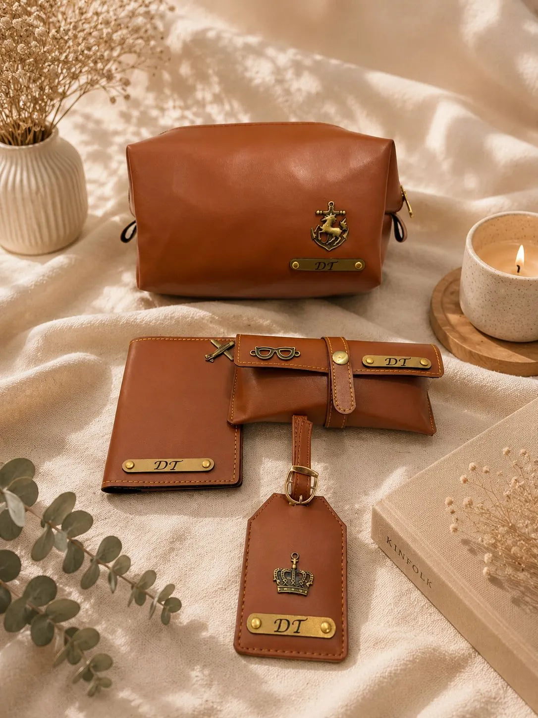 A coordinated set of brown leather accessories arranged on a cream fabric surface, including a zip pouch, wallet, card holder, and luggage tag, each with gold-tone hardware and a visible 'DT' mark. Decorative dried flowers, a candle, and a book are placed around the items.