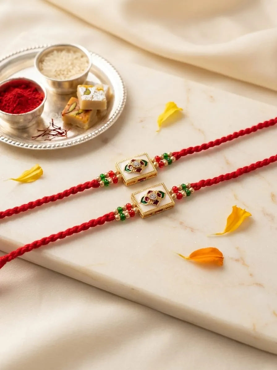 Two red braided Rakhi cords laid diagonally on a cream marble slab, each adorned with a gold-toned rectangular charm featuring a painted portrait and small green and red bead accents. A silver tray with red powder, rice, and sweets sits in the upper left, with soft cream fabric draped around the scene.