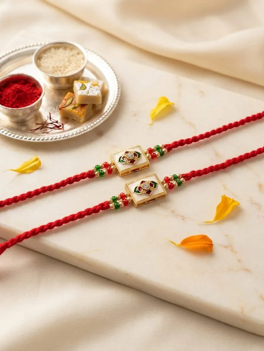 Two red braided Rakhi cords laid diagonally on a cream marble slab, each adorned with a gold-toned rectangular charm featuring a painted portrait and small green and red bead accents. A silver tray with red powder, rice, and sweets sits in the upper left, with soft cream fabric draped around the scene.