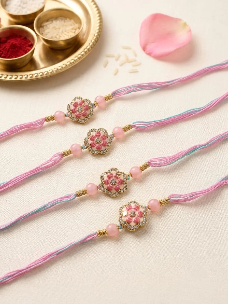 Four pink and blue braided silk Rakhi bracelets with floral beadwork and gold-tone details, arranged diagonally on a light beige surface with a gold tray, rice, and a pink petal in the background.