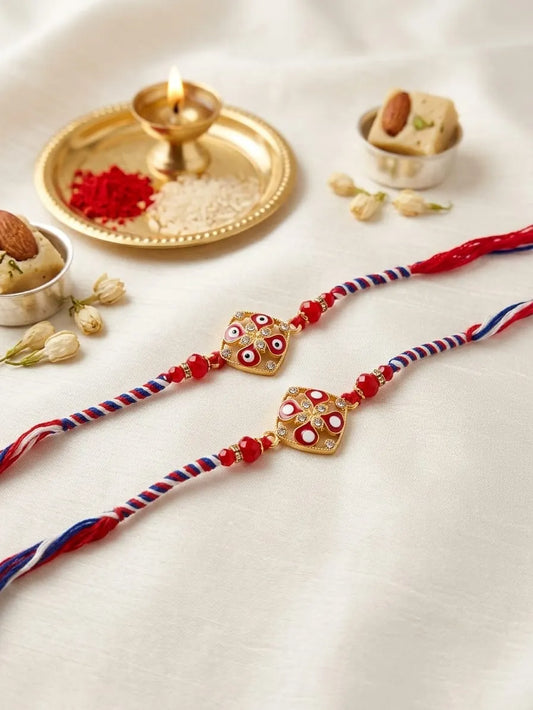 Two traditional Indian Rakhi strings with red, white, and blue braided cords and gold heart-shaped charms decorated with red and white eye motifs, displayed on a white fabric background with festive props including a brass diya, red powder, and sweets.