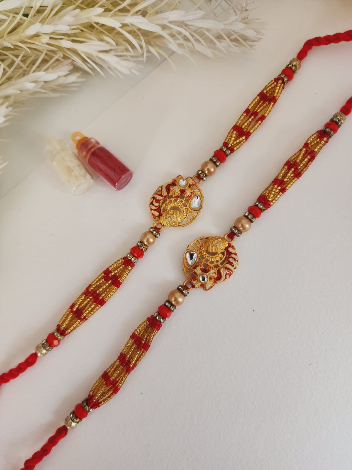 A Hindu woman's rakhi with intricate gold, green, and red beadwork, and a floral design, displayed alongside a small red bottle and a card.