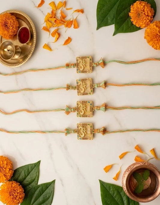Four traditional Indian Rakhi string bracelets with gold-toned Om pendants are arranged on a white marble surface with festive flat-lay props including marigold flowers, green leaves, and copper vessels.