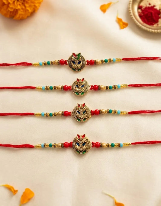 Four red string rakhi bracelets with gold-tone owl pendants and colorful bead accents, arranged vertically on a cream fabric background with decorative orange and red floral elements.