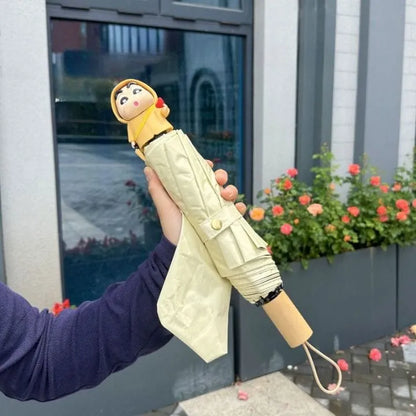A hand holds a closed umbrella outdoors. The umbrella has a cartoon character head at the top, a pale yellow body, and a light wood-toned handle.