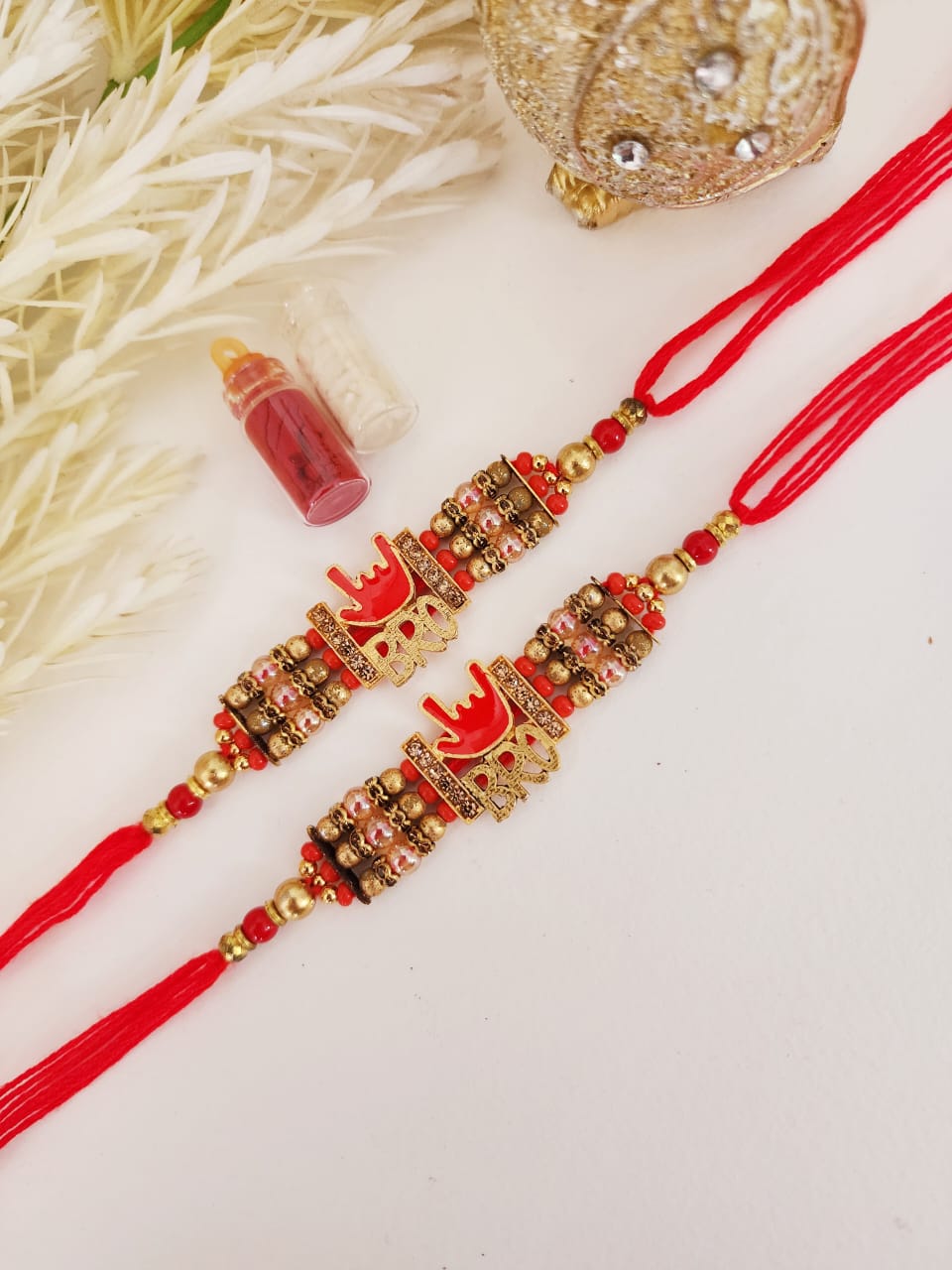 Two red rakhis with gold, silver, and multicolored beadwork, displayed alongside small containers and a decorative object.