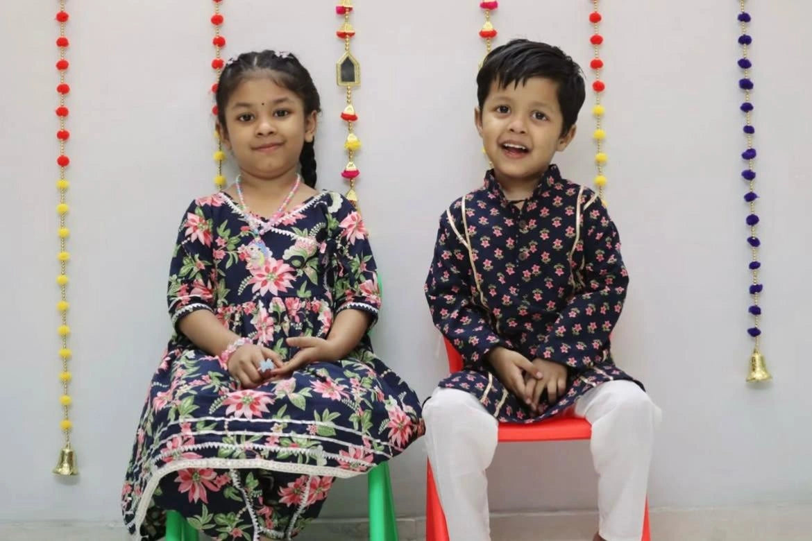 Two children in matching traditional outfits sitting on colorful chairs with decorative hanging beads.