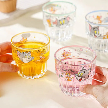 Two glass cups with cartoon characters, one yellow and one pink, held by hands on a light surface.