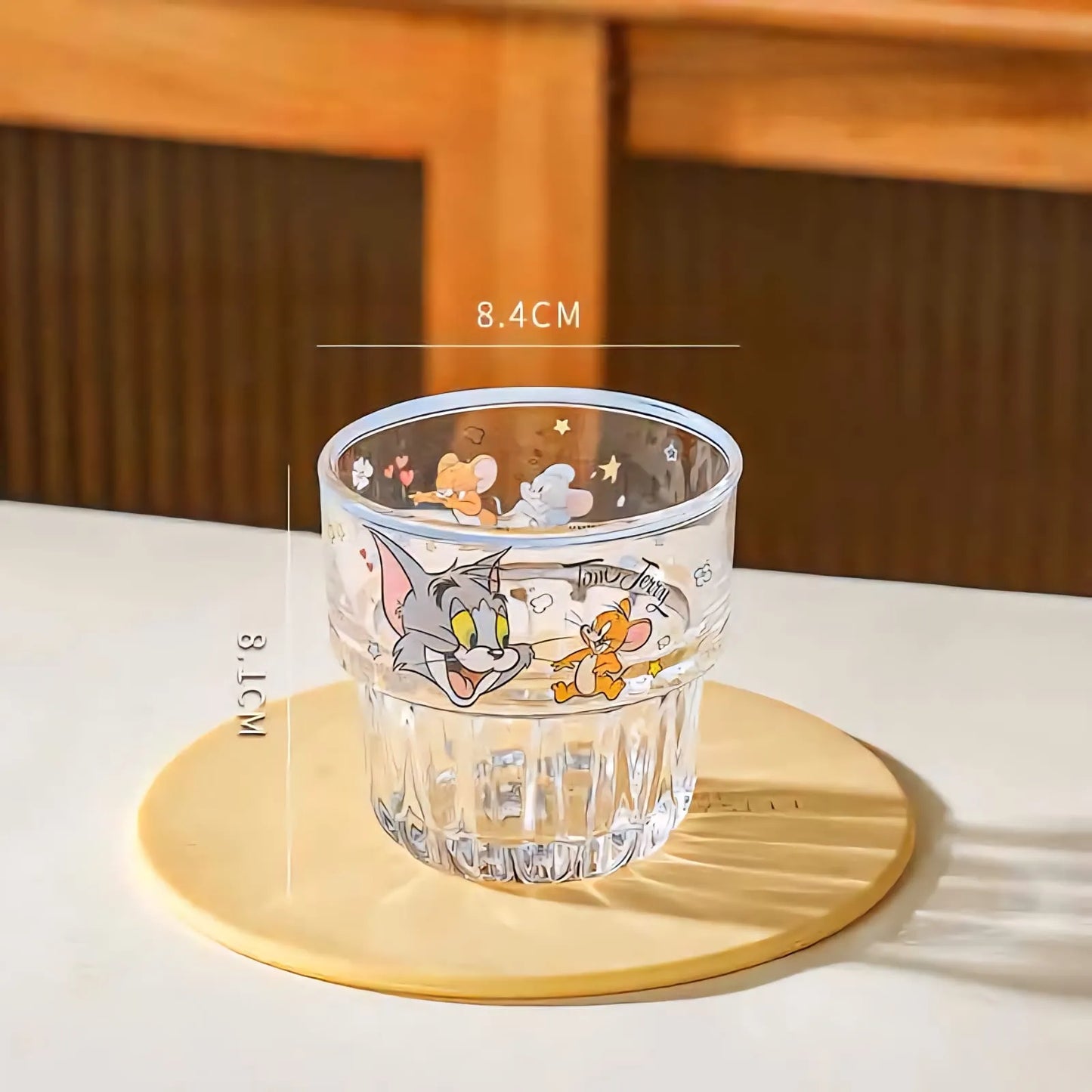 A clear glass tumbler with cartoon graphics of Tom and Jerry, placed on a round beige coaster on a white table. White measurement lines and text overlay indicate dimensions of 8.