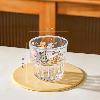 A clear glass tumbler with cartoon graphics of Tom and Jerry, placed on a round beige coaster on a white table. White measurement lines and text overlay indicate dimensions of 8.